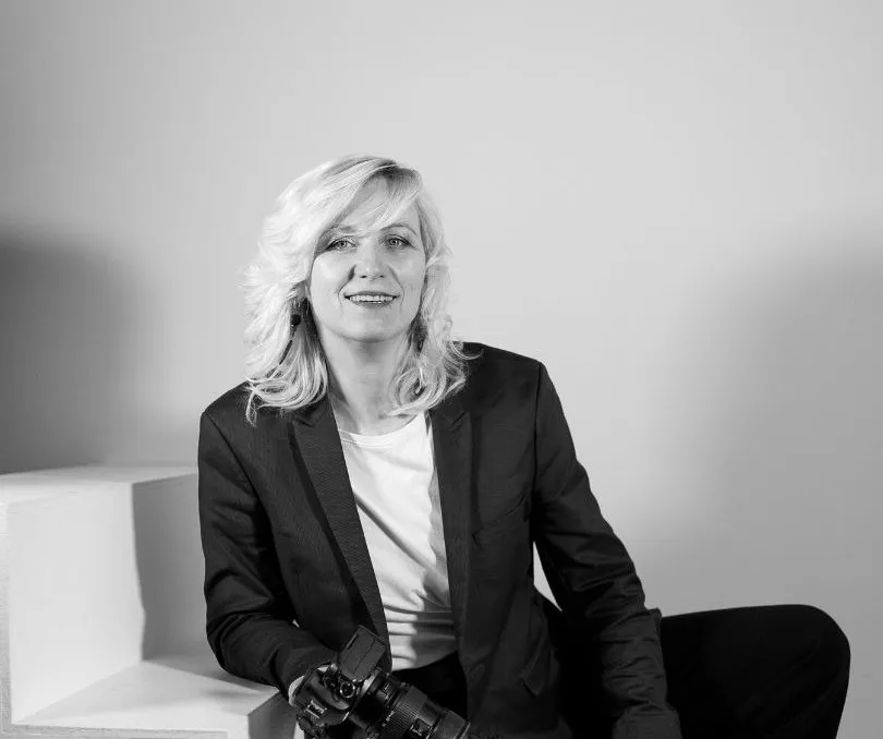 Vineta Jaunzeme melnbaltā portretā sēž uz baltām kāpnēm ar kameru rokās